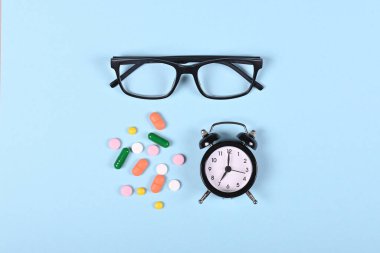 Tıbbi haplar, gözlükler ve mavi arka planda saat, düz yatış, yukarıdan görüntü. Uyuşturucu konseptini kullanma zamanı.