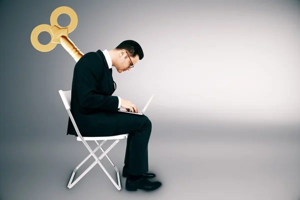 Businessman with wind up key on his back sitting on chair and using laptop computer on light grey background. Manipulation concept — Stock Photo, Image