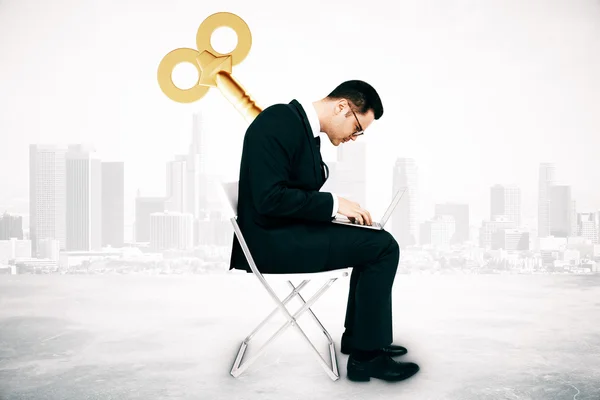 Geschäftsmann mit Klappschlüssel auf dem Rücken, auf einem Stuhl sitzend und mit Laptop im Hintergrund. Manipulationskonzept — Stockfoto