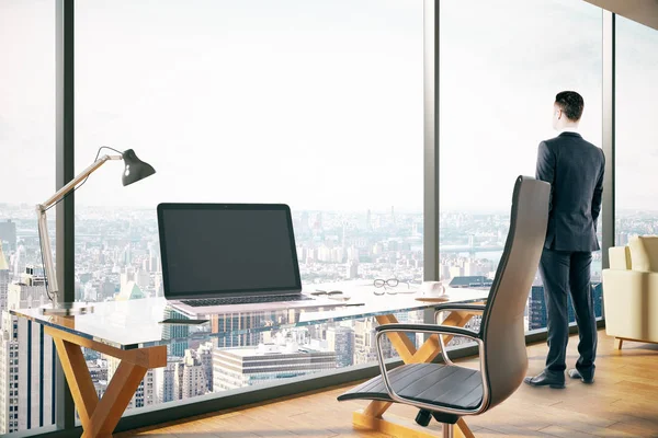 Seitenansicht eines jungen Mannes, der aus dem Fenster in ein modernes Bürointerieur mit leerem Laptop und Stadtblick blickt. mock up, 3D-Darstellung — Stockfoto