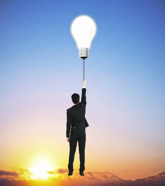 Back view of young businessman flying with abstract light bulb balloon above sunset. Idea concept — Stock Photo, Image