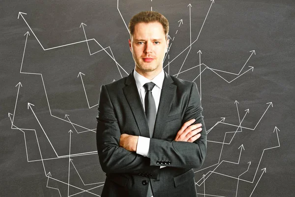 Portrait of confident young businessman with folded arms on chalkboard background with numerous arrows. Direction concept — Stock Photo, Image