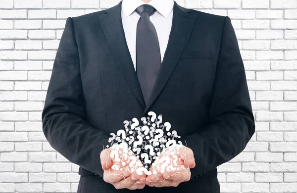 Businessman holding pile of drawn question marks on white brick background. Confusion concept — Stock Photo, Image