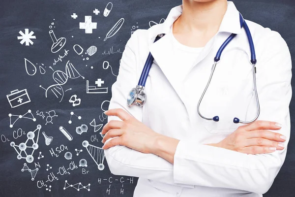 Junge Ärztin mit Stethoskop und verschränkten Armen, die auf Kreidetafel-Hintergrund mit Zeichnungen stehen. Medizinkonzept — Stockfoto