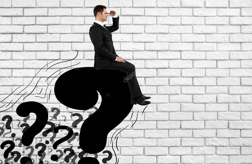 Side view of young businessman sitting on drawn question marks and looking into the distance on brick background with copy space. FAQ concept 