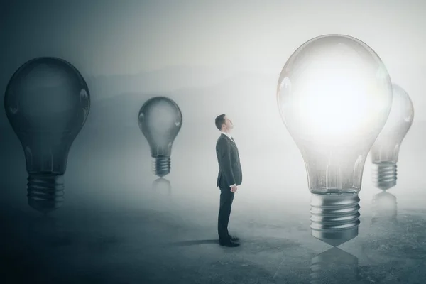 Вид сбоку на молодого бизнесмена, смотрящего на светящиеся лампы на туманном сером фоне. Концепция исследования. 3D рендеринг — стоковое фото