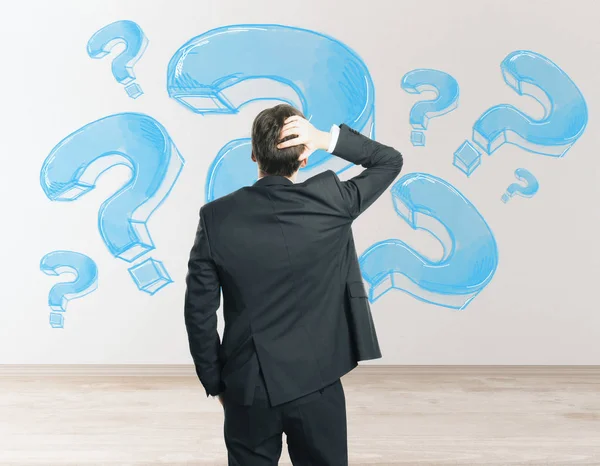 Back view of stressed young businessman looking at concrete wall with drawn question marks in interior. Worry concept — Stock Photo, Image