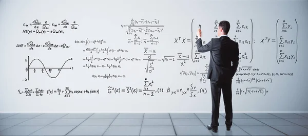 Vue arrière de jeune homme d'affaires écrivant des formules mathématiques sur le mur de béton à l'intérieur. Concept scientifique — Photo