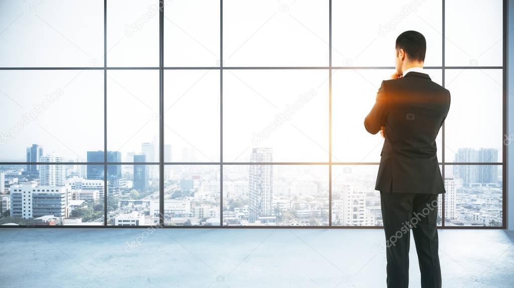 Back view of thoughtful young businessman standing in modern concrete office with panoramic city view and daylight. Research concept. 3D Rendering 