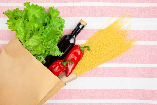 Paper bag with spaghetti, oil, pepper and salad on tablecloth.