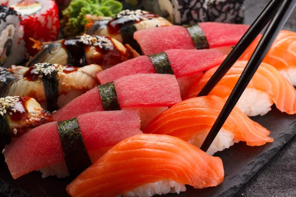 Japanese cuisine. Sushi in black chopsticks on a stone plate and black concrete background. — Stock Photo, Image