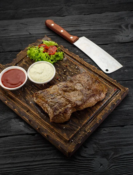 Bife grelhado com molhos numa tábua. Mesa de madeira escura . — Fotografia de Stock