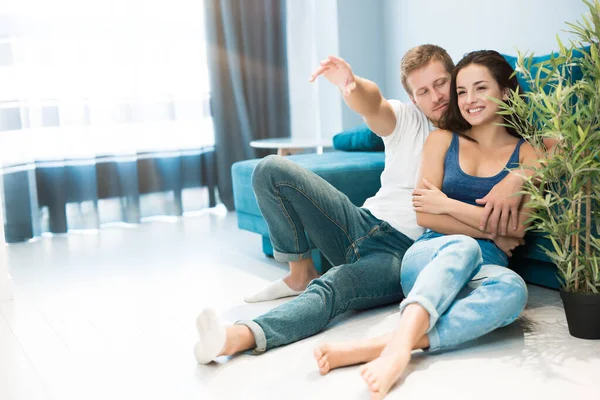 Jong gelukkig paar zitten op de vloer knuffelen thuis in de woonkamer man tonen aan vriendin iets grappigs met zijn vinger familie reünie — Stockfoto