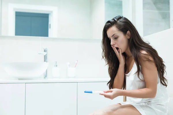 Giovane bella donna bruna siede sul water tenendo il test di gravidanza cercando sorpresi momenti imprevedibili di vita — Foto Stock