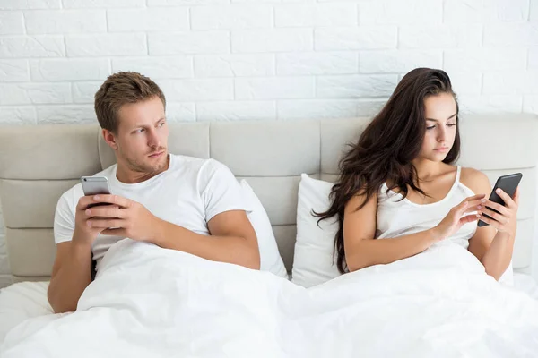 Young couple handsome husband and his beautiful wife just woke up in bed checking their smartphones early in the morning man looks suspective social media addiction — Stock Photo, Image