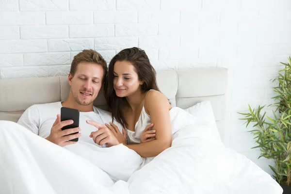 Young couple handsome husband and his beautiful wife just woke up in bed watching photoes on smartphone early in the morning social media addiction — Stock Photo, Image