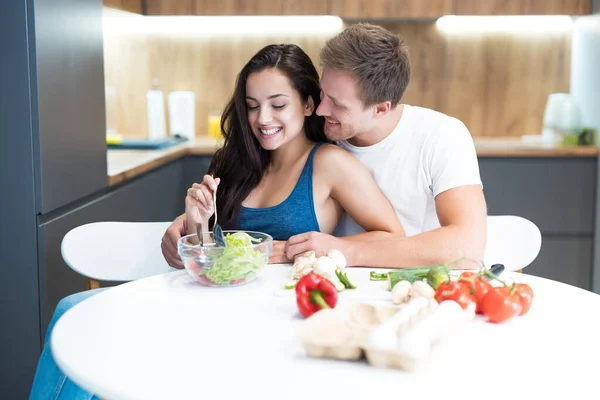 Jong paar koken ontbijt in de keuken samen man knuffelt zijn geliefde vrouw terwijl ze mengt de salade familie tradities — Stockfoto