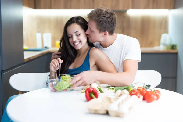 Jong paar koken ontbijt in de keuken samen man knuffels en kust zijn geliefde vrouw terwijl ze mengt de salade familie tradities — Stockfoto