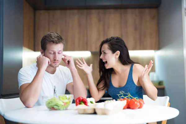 Fiatal pár veszekedés közben főzés reggeli a konyhában feleség kiabál hangosan férje ő becsukja a fülét ujjaival néz bosszús családi veszekedés — Stock Fotó