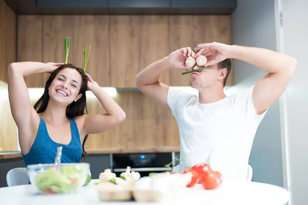 Jovem casal engraçado cozinhar café da manhã na cozinha homem fecha os olhos com cogumelos, enquanto sua amada esposa segura cebola verde como orelhas rindo juntos — Fotografia de Stock
