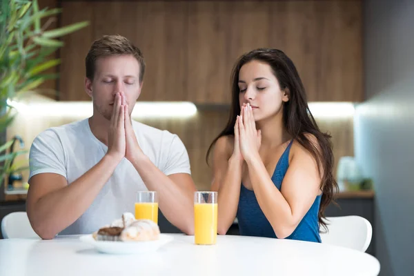 Young couple saying prayer before start eating their breakfast in the kitchen family traditions — Stock Photo, Image