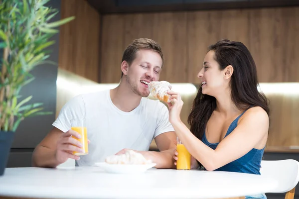 Jong mooi paar eten croissant en drinken sinaasappelsap voor het ontbijt in de keuken vrouw voedt haar man begin van grote dag — Stockfoto