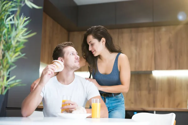 Unga vackra par äter croissant och dricka apelsinjuice till frukost i köket kvinnan står bakom sin man kramar honom bra början på dagen — Stockfoto