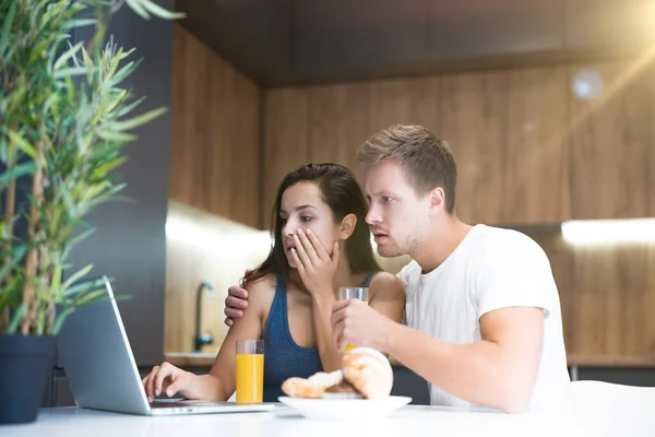 Joven pareja guapo hombre y hermosa mujer se ve sorprendido viendo en la pantalla del ordenador portátil mientras toma croissants con jugo de naranja para el desayuno en la cocina — Foto de Stock