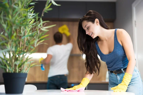 Young man with detergent spray wiping dust off from kitchen cabinet with rag meanwhile smiling woman wiping kitchen table in yellow gloves doing cleaning together — Stock Photo, Image