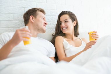 Mutlu, genç, gülümseyen bir çift yakışıklı adam ve güzel bir kadın sabahın köründe parlak yatak odasında uzanmış portakal suyu içiyorlar.