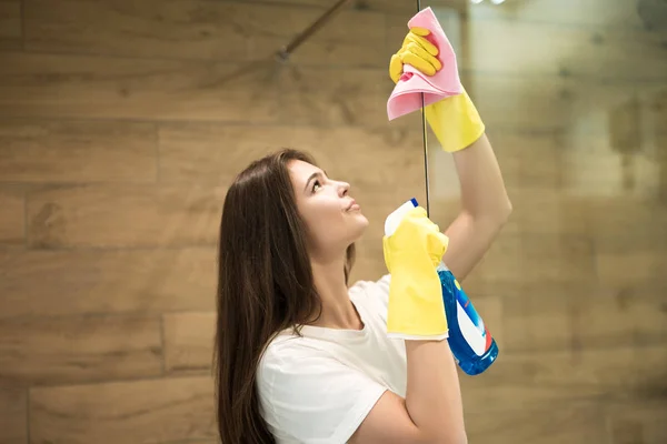 Beautiful young woman in yellow gloves spraying detergent spray while wiping dust off from the shower door with a rag in the WC — Stock Photo, Image