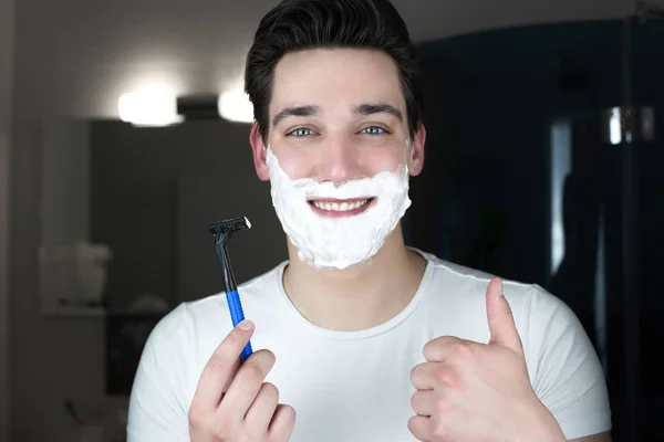 Young handsome smiling man with foam on his holding razor in his hand showing like sign in bathroom in the morning l everyday routine — Stock Photo, Image
