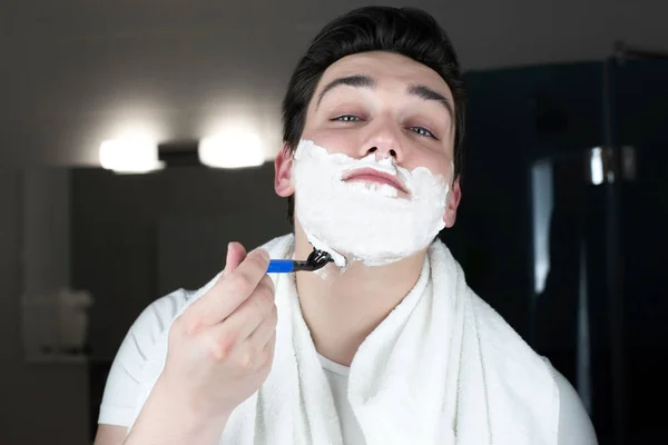 young handsome man with foam on his face shaving with razor in bathroom in the morning everyday routine