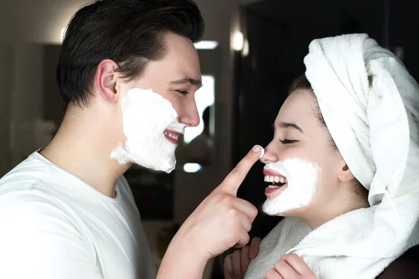 Funny couple handsome man applying jokingly foam on beautiful woman's nose having fun both with shaving foam on their faces in bathroom laughing humor moment — Stock Photo, Image