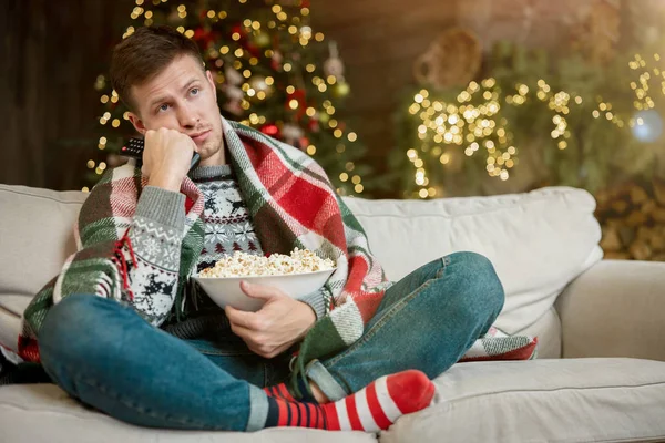 Joven hombre guapo envuelto en cuadros viendo la televisión comiendo palomitas de maíz en el sofá en la habitación decorada para celebrar la Navidad de año nuevo solo estado de ánimo melancólico —  Fotos de Stock