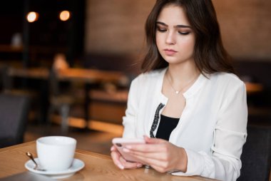 Genç ve güzel bir kadın kafeteryada sıcak kahve içerken akıllı telefonunu kontrol ediyor.