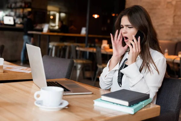 Joven hermosa mujer que trabaja en su computadora portátil mirando enojado durante la conversación telefónica mientras bebe café caliente en la cafetería multitarea moderna mujer de negocios — Foto de Stock