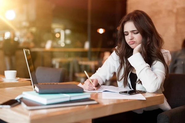 young woman in white jacket working in her laptop taking notes in her planner looking annoyed while drinking hot coffee in cafe multitasking modern businesswoman
