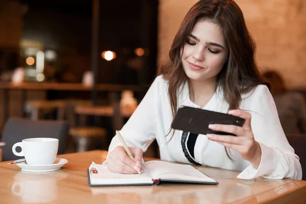 Νεαρή όμορφη γυναίκα κρατώντας σημειώσεις για τον σχεδιαστή της χρησιμοποιώντας το smartphone της πίνοντας ζεστό καφέ στο cafe multitasking σύγχρονη επιχειρηματίας — Φωτογραφία Αρχείου