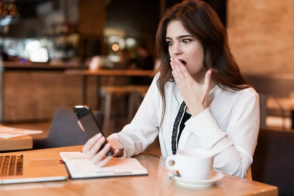 Joven hermosa mujer con el teléfono en su mano trabajando fuera de la oficina en el ordenador portátil se ve sorprendido de recibir malas noticias beber café caliente en la cafetería moderna mujer de negocios — Foto de Stock