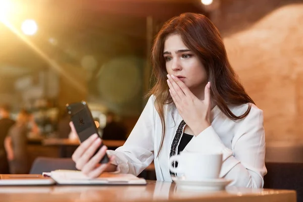 Joven mujer molesta con el teléfono en su mano trabajando fuera de la oficina en el ordenador portátil se ve sorprendido de recibir malas noticias beber café caliente en la cafetería moderna mujer de negocios — Foto de Stock