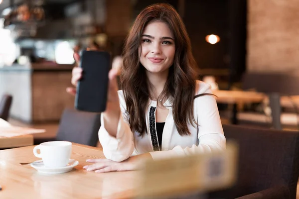 Νέα χαμογελαστή γυναίκα δείχνει το smartphone της κατά τη διάρκεια του διαλείμματος γεύμα πίνοντας ζεστό καφέ στο μοντέρνο καφέ αναζητούν ευτυχισμένη σύγχρονη επιχειρηματίας — Φωτογραφία Αρχείου