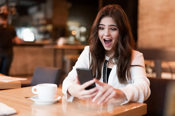 Joven hermosa mujer en chaqueta blanca con estilo con teléfono inteligente en sus manos mirando sorprendido durante el descanso de café en la cafetería moderna mujer de negocios — Foto de Stock