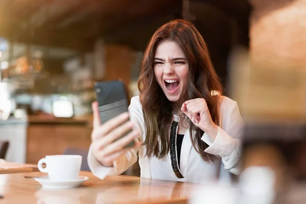 Νεαρή όμορφη γυναίκα σε λευκό κομψό σακάκι με smartphone στα χέρια της αναζητούν νικηφόρα λήψη φανταστική είδηση κατά τη διάρκεια του διαλείμματος καφέ στο καφενείο σύγχρονη επιχειρηματίας — Φωτογραφία Αρχείου