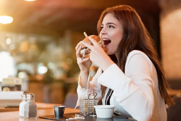 Młoda piękna kobieta w białej stylowej bluzce wygląda głodny jedzenie mięsa burger z frytkami na obiad w modnej kawiarni jedzenie na zewnątrz — Zdjęcie stockowe