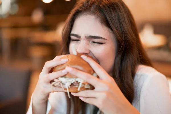 Joven hambrienta mujer en blanco elegante blusa morder con apetito carne fresca hamburguesa durante el almuerzo en la moda restaurante comer al aire libre —  Fotos de Stock