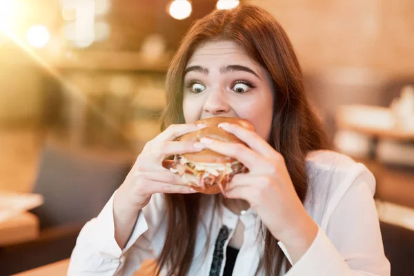 Wanita muda dalam blus putih bergaya menggigit dengan nafsu makan daging segar lezat burger selama makan siang di kafe trendi tampak sangat lapar makan di luar — Stok Foto
