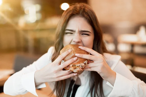 Wanita muda dalam blus putih bergaya menggigit dengan burger segar nafsu makan selama makan siang di kafe trendi merasa lapar utama makan di luar — Stok Foto