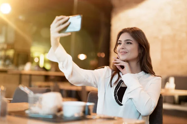Νεαρή όμορφη γυναίκα σε λευκό κομψό μπλούζα φαίνεται ωραίο, ενώ τη λήψη selfie πριν από το κρέας burger με πατάτες για μεσημεριανό γεύμα σε μοντέρνο καφέ τρώει έξω — Φωτογραφία Αρχείου