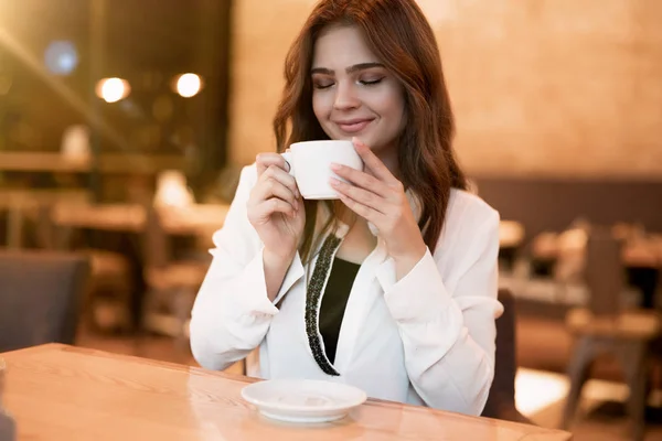 young beautiful woman in white stylish blouse drinking hot coffee after workday sitting in trendy cafe modern businesswoman multitasking